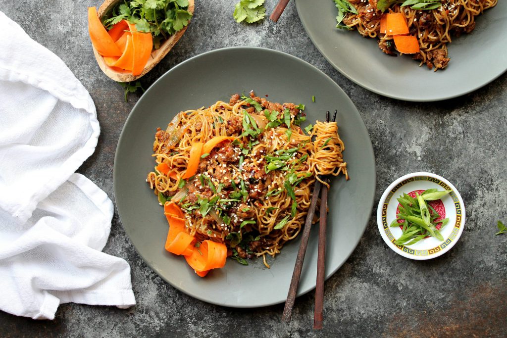 A close-up of crispy ginger ‘beef’ ramen stir-fry in a bowl, garnished with pickled carrots, chili oil, and sesame seeds.