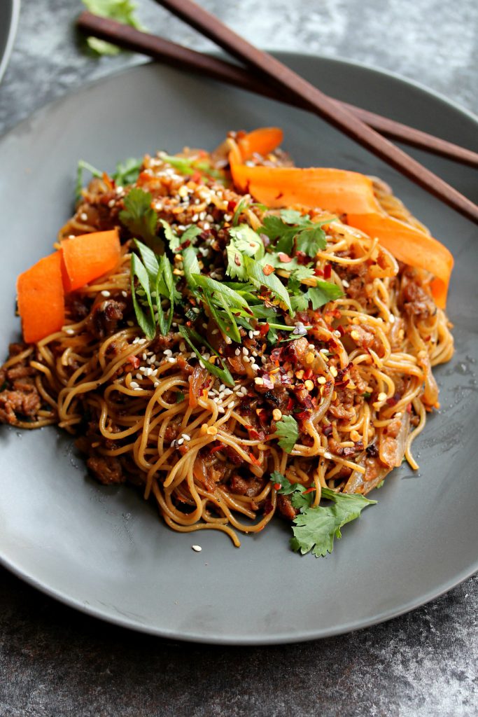 A close-up of crispy ginger ‘beef’ ramen stir-fry in a bowl, garnished with pickled carrots, chili oil, and sesame seeds.