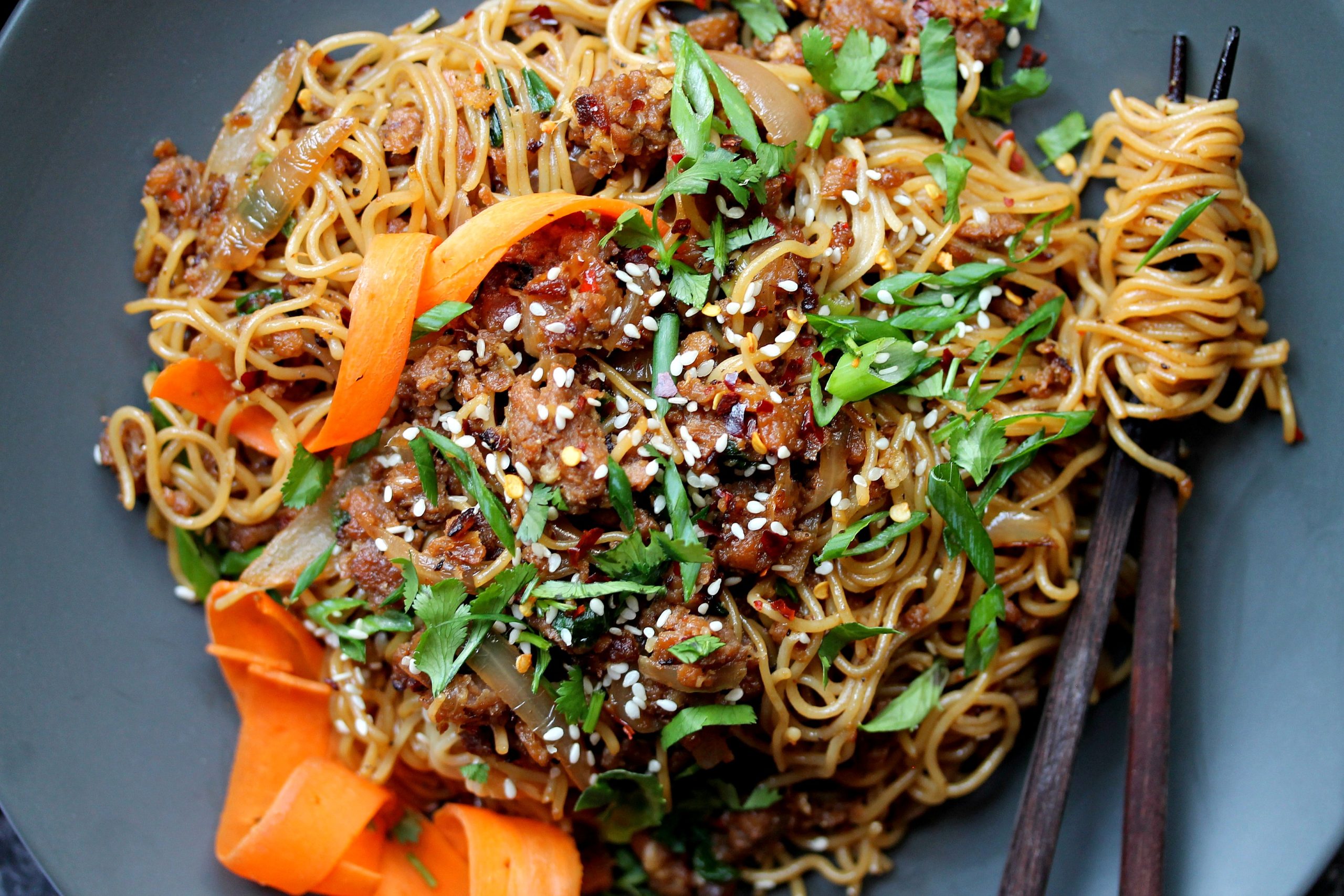 A close-up of crispy ginger ‘beef’ ramen stir-fry in a bowl, garnished with pickled carrots, chili oil, and sesame seeds.