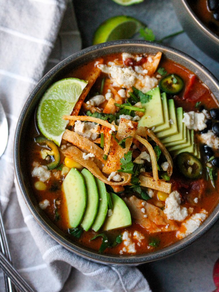 Bowl of hearty vegan tortilla soup garnished with crispy tortilla strips, sliced avocado, fresh cilantro, and a lime wedge.