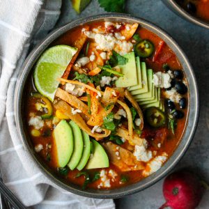 Bowl of hearty vegan tortilla soup garnished with crispy tortilla strips, sliced avocado, fresh cilantro, and a lime wedge.