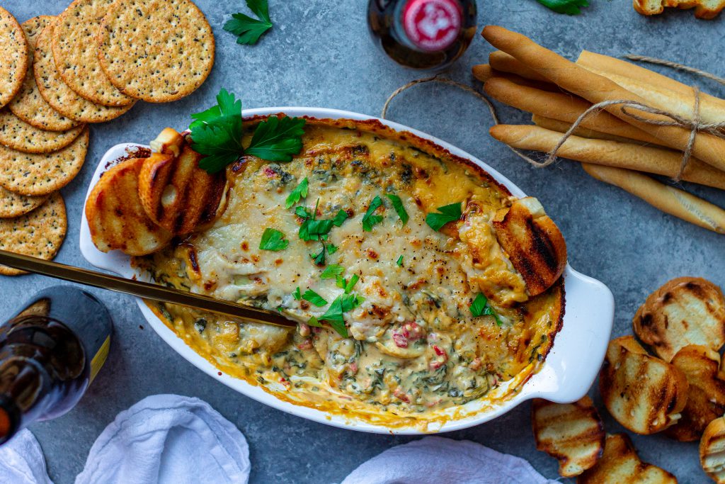 A creamy Vegan Spinach Artichoke Dip in a baking dish, topped with melted vegan parmesan and garnished with fresh parsley, surrounded by pita chips, toasted baguette slices, and veggies.