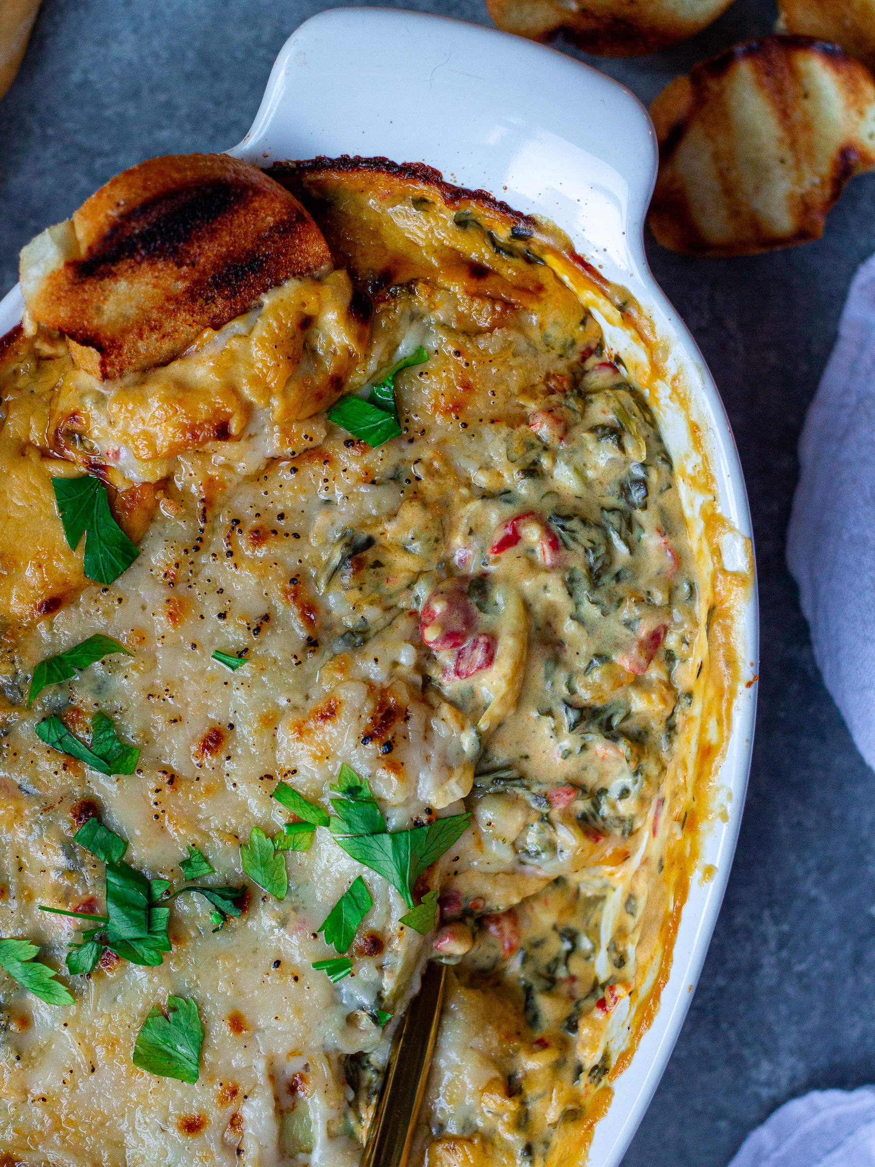 A creamy Vegan Spinach Artichoke Dip in a baking dish, topped with melted vegan parmesan and garnished with fresh parsley, surrounded by pita chips, toasted baguette slices, and veggies.