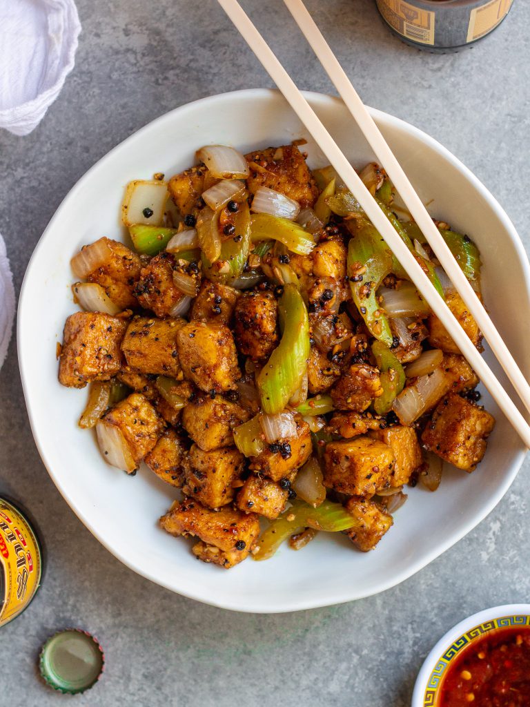 Close-up of crispy black pepper tofu with sautéed onion and crisp celery tossed in a glossy sauce.