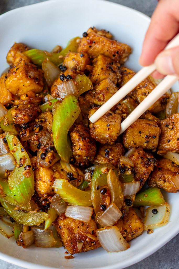 Close-up of crispy black pepper tofu with sautéed onion and crisp celery tossed in a glossy sauce.