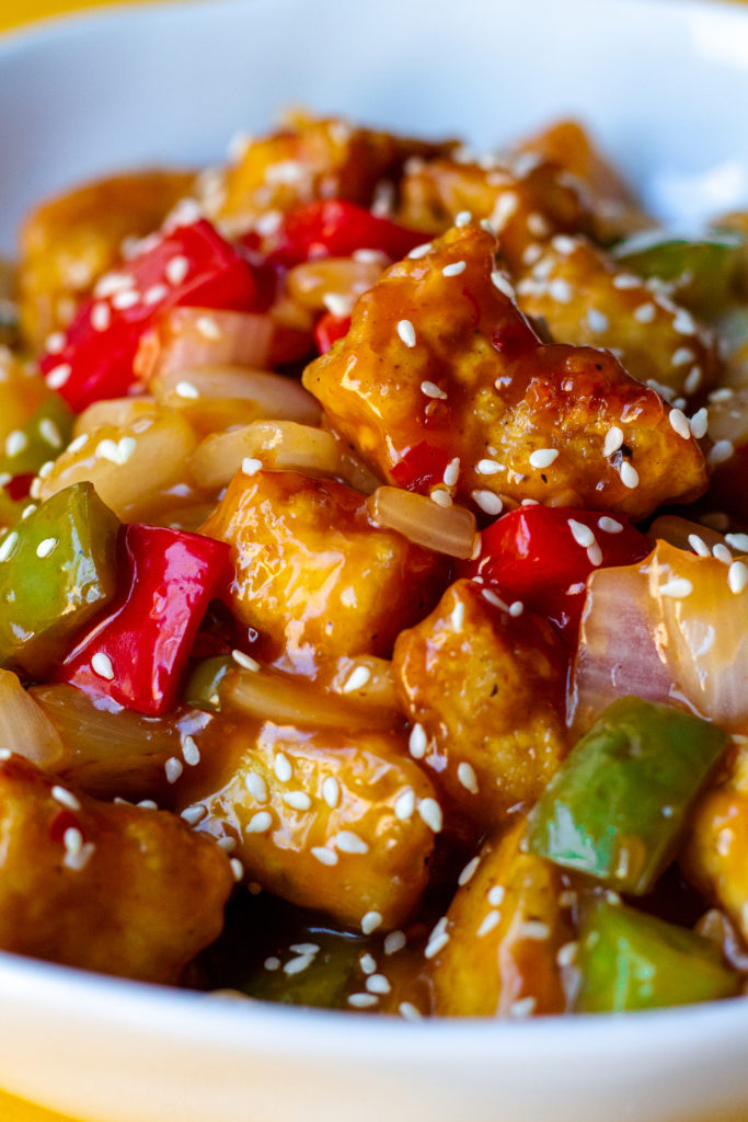 A plate of crispy sweet and sour tofu with bell peppers and pineapple over white rice garnished with toasted sesame seeds and thinly sliced green onion.