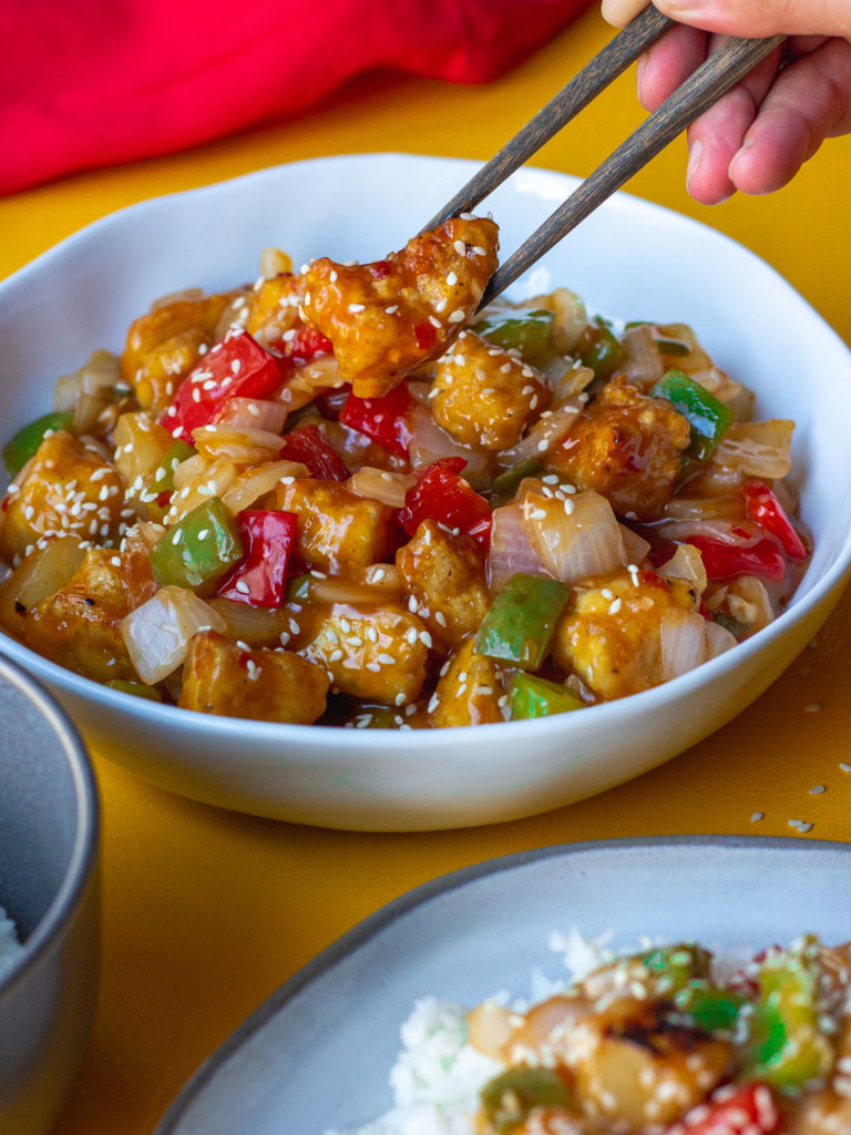 A plate of crispy sweet and sour tofu with bell peppers and pineapple over white rice garnished with toasted sesame seeds and thinly sliced green onion.