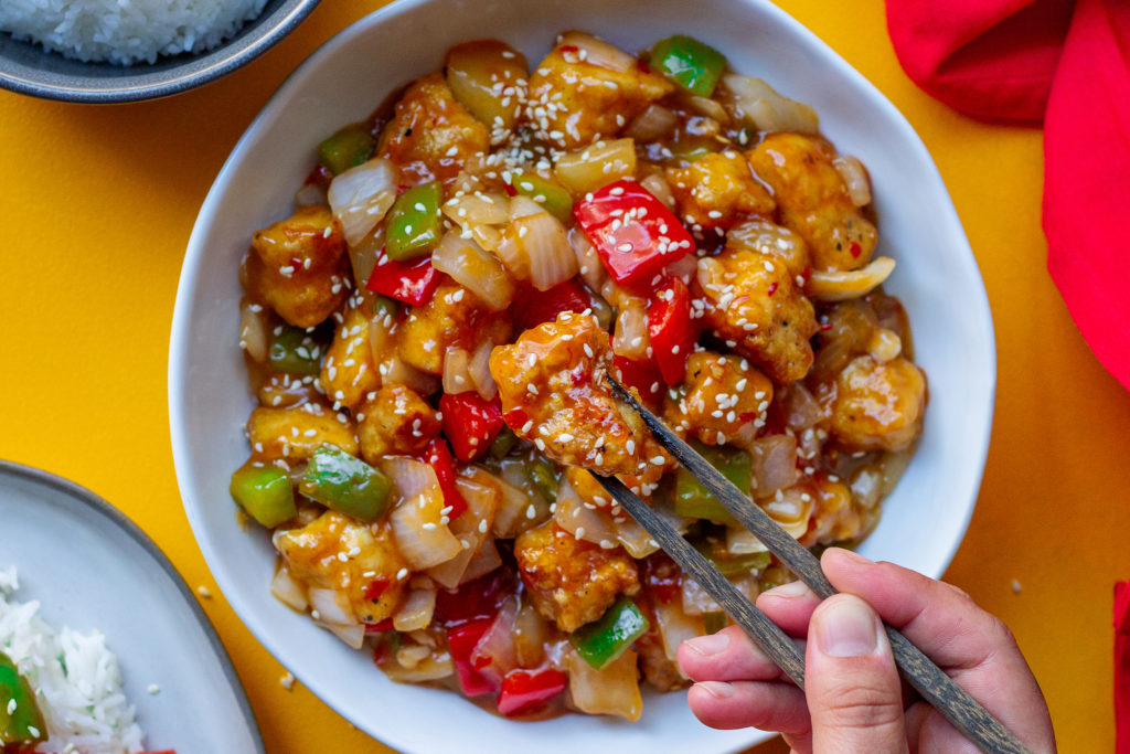 A plate of crispy sweet and sour tofu with bell peppers and pineapple over white rice garnished with toasted sesame seeds and thinly sliced green onion.