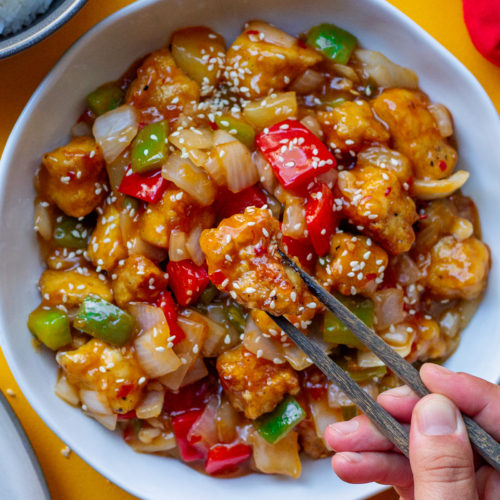 A plate of crispy sweet and sour tofu with bell peppers and pineapple over white rice garnished with toasted sesame seeds and thinly sliced green onion.