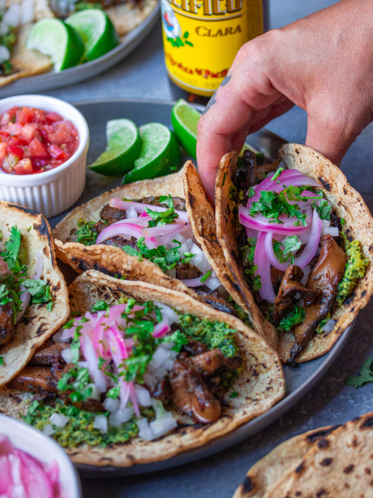A plate of finished Vegan Asada Mushroom Tacos garnished with fresh lime wedges, cilantro, and hot sauce on the side.
