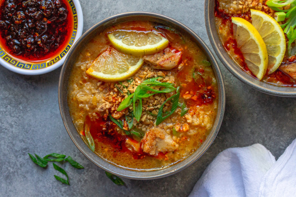 Bowl of creamy and comforting vegan arroz caldo garnished with chili oil, fried garlic, green onions, and lemon wedges.