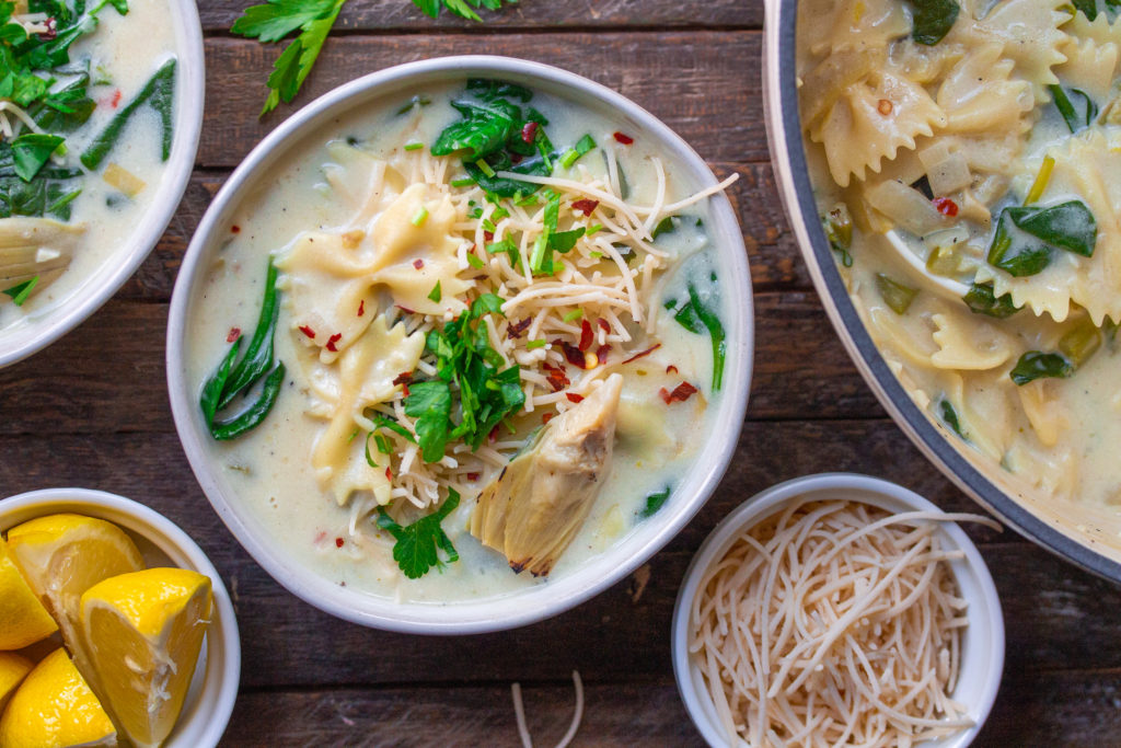 A pot of creamy soup with noodles, spinach, and artichoke hearts on a stovetop.