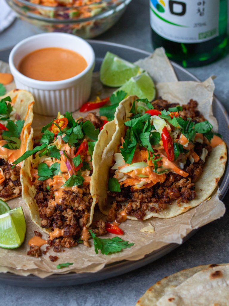 A photo of the assembled tacos, with warm tortillas filled with the bulgogi-inspired tofu filling, drizzled with gochujang sauce, topped with slaw and garnished with fresh cilantro and sliced Thai chili, with a lime wedge on the side.