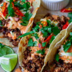 A photo of the assembled tacos, with warm tortillas filled with the bulgogi-inspired tofu filling, drizzled with gochujang sauce, topped with slaw and garnished with fresh cilantro and sliced Thai chili, with a lime wedge on the side.