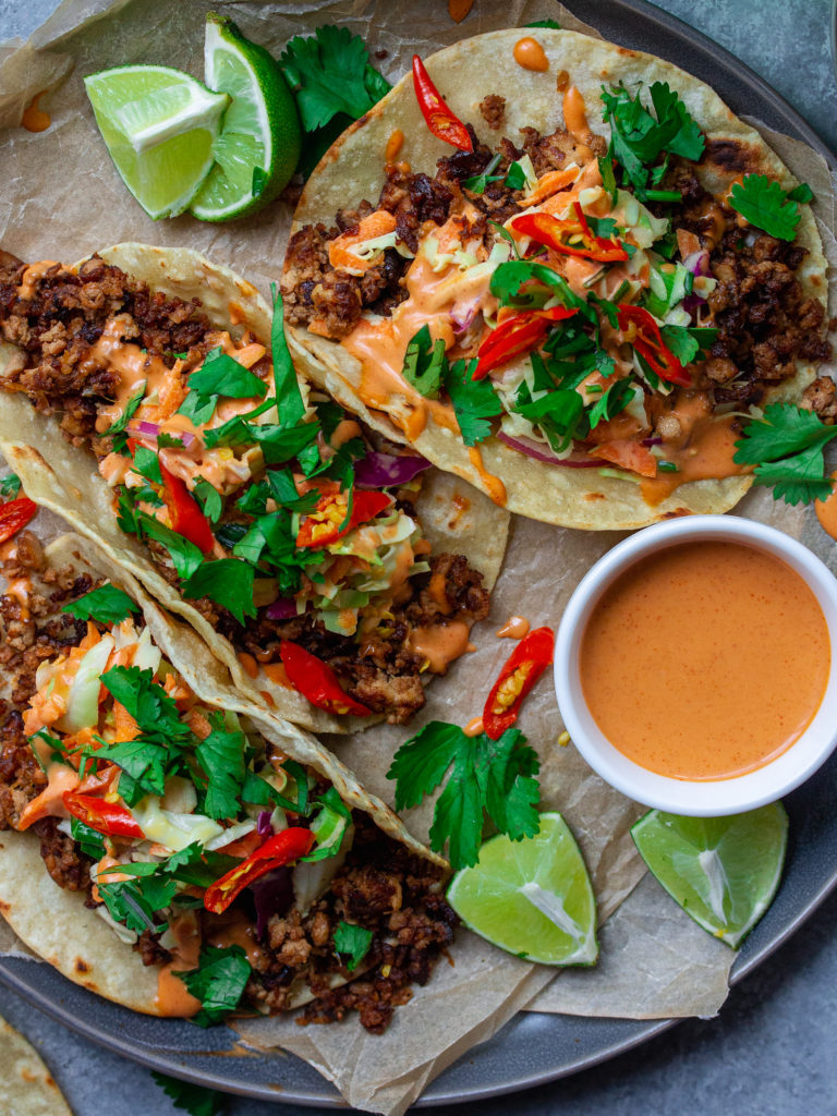 A photo of the assembled tacos, with warm tortillas filled with the bulgogi-inspired tofu filling, drizzled with gochujang sauce, topped with slaw and garnished with fresh cilantro and sliced Thai chili, with a lime wedge on the side.