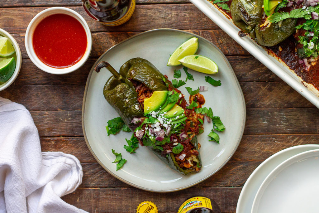 A plate of beautifully roasted poblano peppers filled with seasoned vegan ground, roasted corn, and rice. Topped with melted vegan cheese and garnished with sliced avocado, cilantro, red onion, and roasted corn.