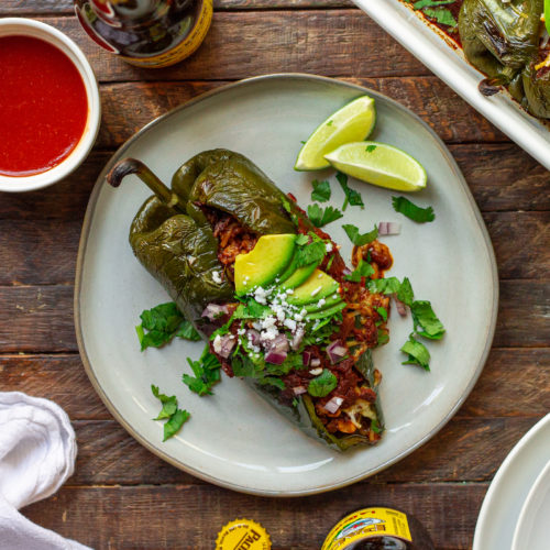 A plate of beautifully roasted poblano peppers filled with seasoned vegan ground, roasted corn, and rice. Topped with melted vegan cheese and garnished with sliced avocado, cilantro, red onion, and roasted corn.
