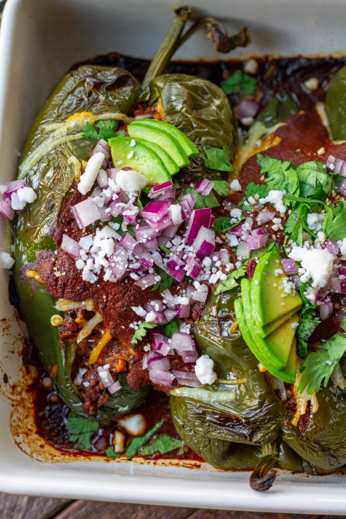 A plate of beautifully roasted poblano peppers filled with seasoned vegan ground, roasted corn, and rice. Topped with melted vegan cheese and garnished with sliced avocado, cilantro, red onion, and roasted corn.