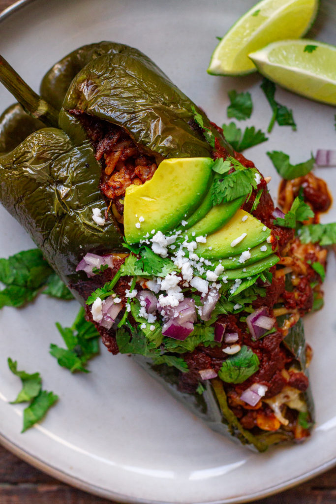 A plate of beautifully roasted poblano peppers filled with seasoned vegan ground, roasted corn, and rice. Topped with melted vegan cheese and garnished with sliced avocado, cilantro, red onion, and roasted corn.