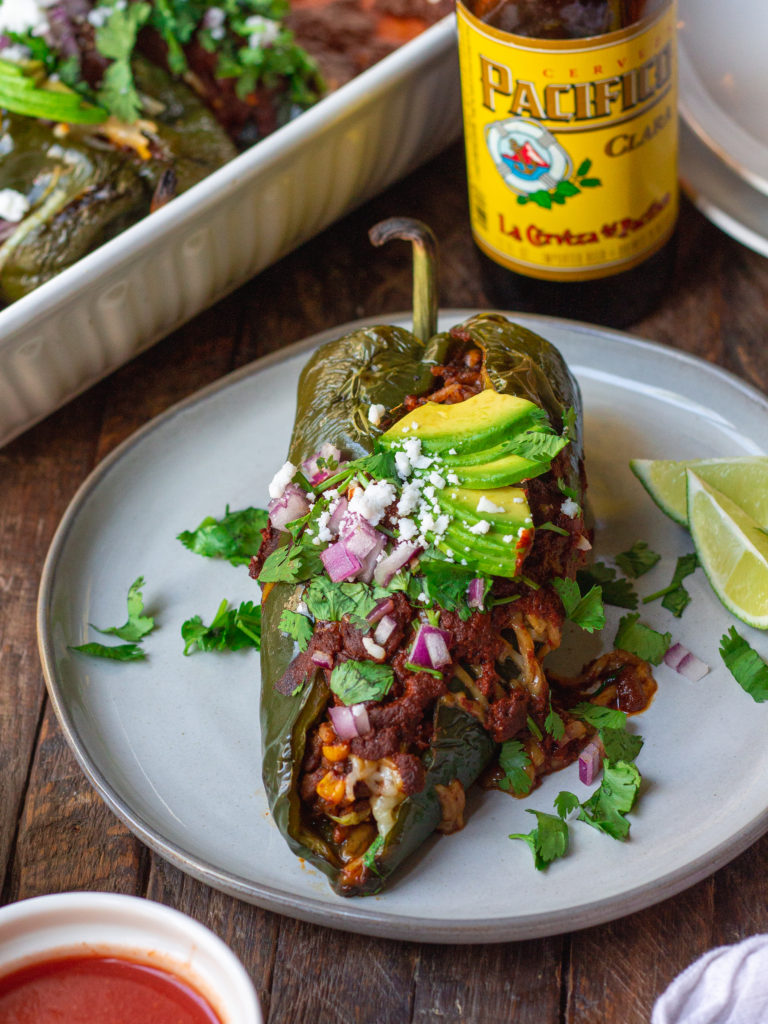 A plate of beautifully roasted poblano peppers filled with seasoned vegan ground, roasted corn, and rice. Topped with melted vegan cheese and garnished with sliced avocado, cilantro, red onion, and roasted corn.