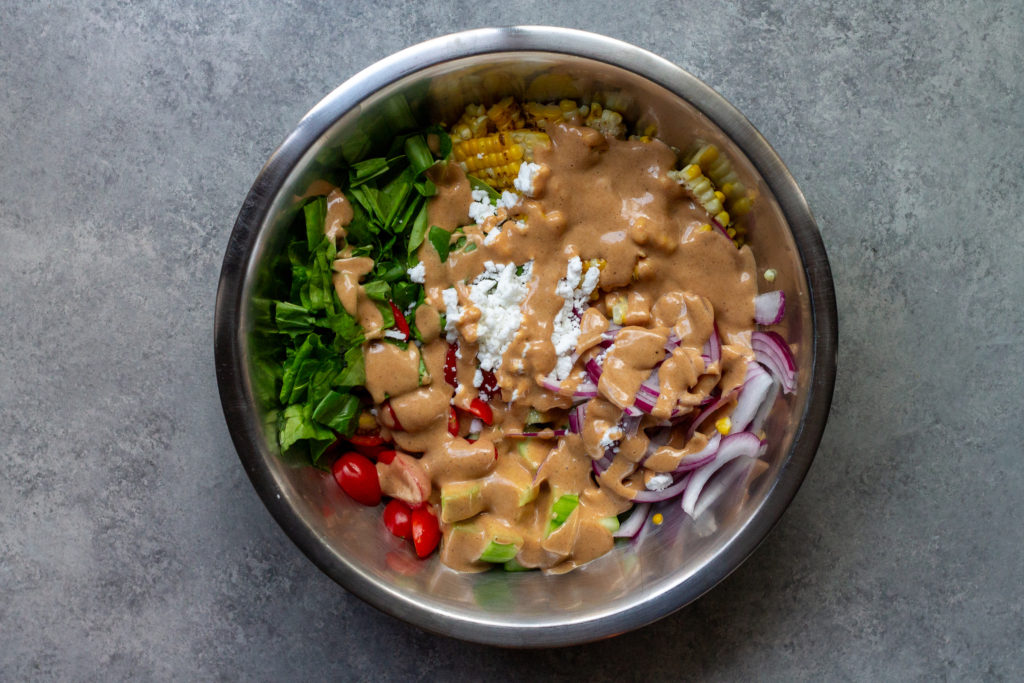 A colorful and vibrant vegan corn, tomato, and bacon salad served in a bowl. The salad is adorned with halved cherry tomatoes, fresh corn kernels, diced cucumber, torn basil leaves, chopped cilantro, and diced avocado. The Bacon Vinaigrette is drizzled over the top, adding a burst of flavor.