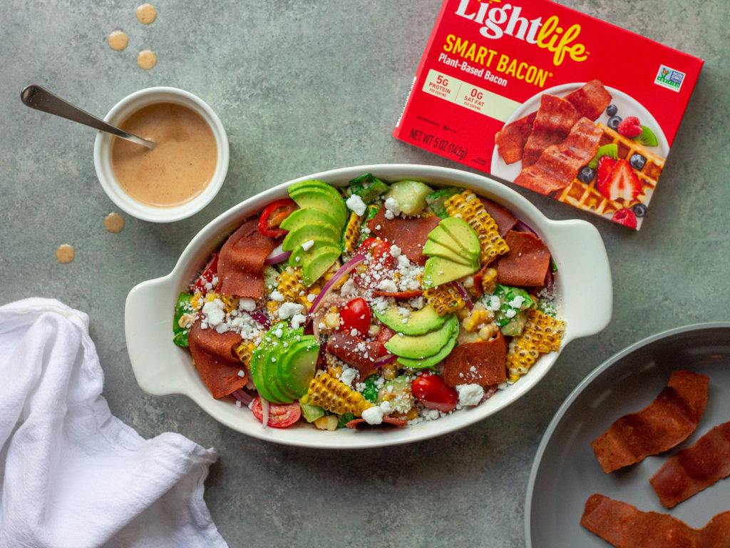 A colorful and vibrant vegan corn, tomato, and bacon salad served in a bowl. The salad is adorned with halved cherry tomatoes, fresh corn kernels, diced cucumber, torn basil leaves, chopped cilantro, and diced avocado. The Bacon Vinaigrette is drizzled over the top, adding a burst of flavor.