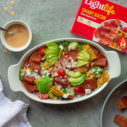 A colorful and vibrant vegan corn, tomato, and bacon salad served in a bowl. The salad is adorned with halved cherry tomatoes, fresh corn kernels, diced cucumber, torn basil leaves, chopped cilantro, and diced avocado. The Bacon Vinaigrette is drizzled over the top, adding a burst of flavor.