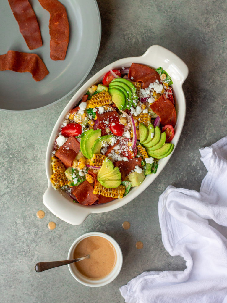 A colorful and vibrant vegan corn, tomato, and bacon salad served in a bowl. The salad is adorned with halved cherry tomatoes, fresh corn kernels, diced cucumber, torn basil leaves, chopped cilantro, and diced avocado. The Bacon Vinaigrette is drizzled over the top, adding a burst of flavor.