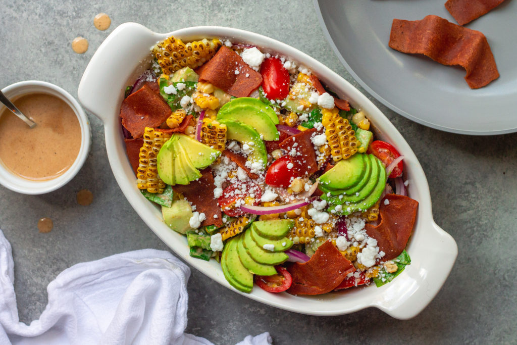 A colorful and vibrant vegan corn, tomato, and bacon salad served in a bowl. The salad is adorned with halved cherry tomatoes, fresh corn kernels, diced cucumber, torn basil leaves, chopped cilantro, and diced avocado. The Bacon Vinaigrette is drizzled over the top, adding a burst of flavor.
