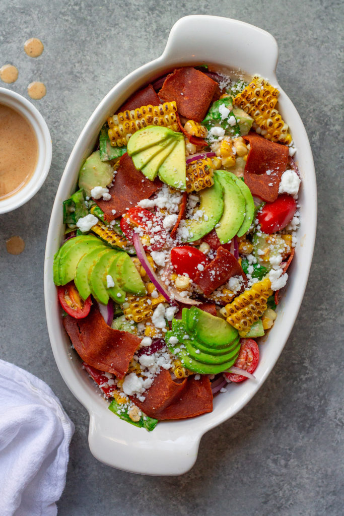 A colorful and vibrant vegan corn, tomato, and bacon salad served in a bowl. The salad is adorned with halved cherry tomatoes, fresh corn kernels, diced cucumber, torn basil leaves, chopped cilantro, and diced avocado. The Bacon Vinaigrette is drizzled over the top, adding a burst of flavor.