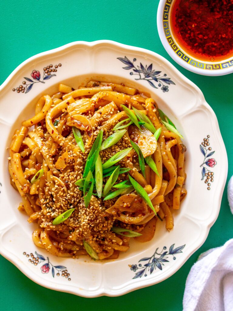 A serving plate with hot chili oil garlic udon noodles, garnished with chopped green onions and toasted sesame seeds.