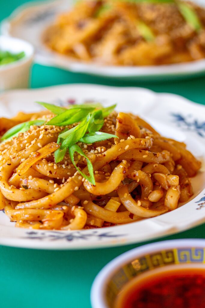 A serving plate with hot chili oil garlic udon noodles, garnished with chopped green onions and toasted sesame seeds.