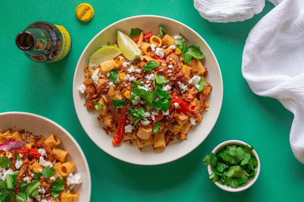 A serving plate showcasing the final dish of Vegan Chipotle Chicken Pasta, topped with vegan feta, bac'n bits, and fresh cilantro, with lime wedges on the side.