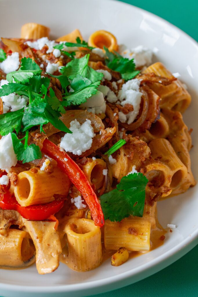 A serving plate showcasing the final dish of Vegan Chipotle Chicken Pasta, topped with vegan feta, bac'n bits, and fresh cilantro, with lime wedges on the side.