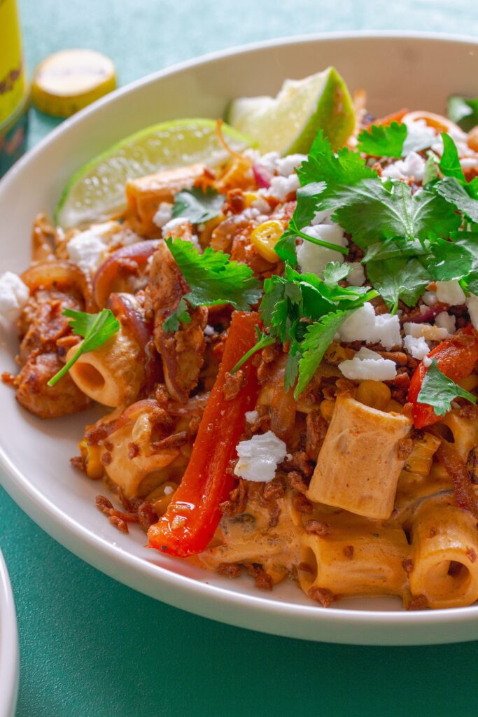 A serving plate showcasing the final dish of Vegan Chipotle Chicken Pasta, topped with vegan feta, bac'n bits, and fresh cilantro, with lime wedges on the side.