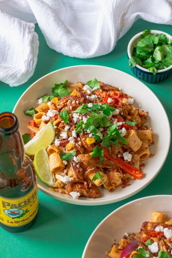 A serving plate showcasing the final dish of Vegan Chipotle Chicken Pasta, topped with vegan feta, bac'n bits, and fresh cilantro, with lime wedges on the side.