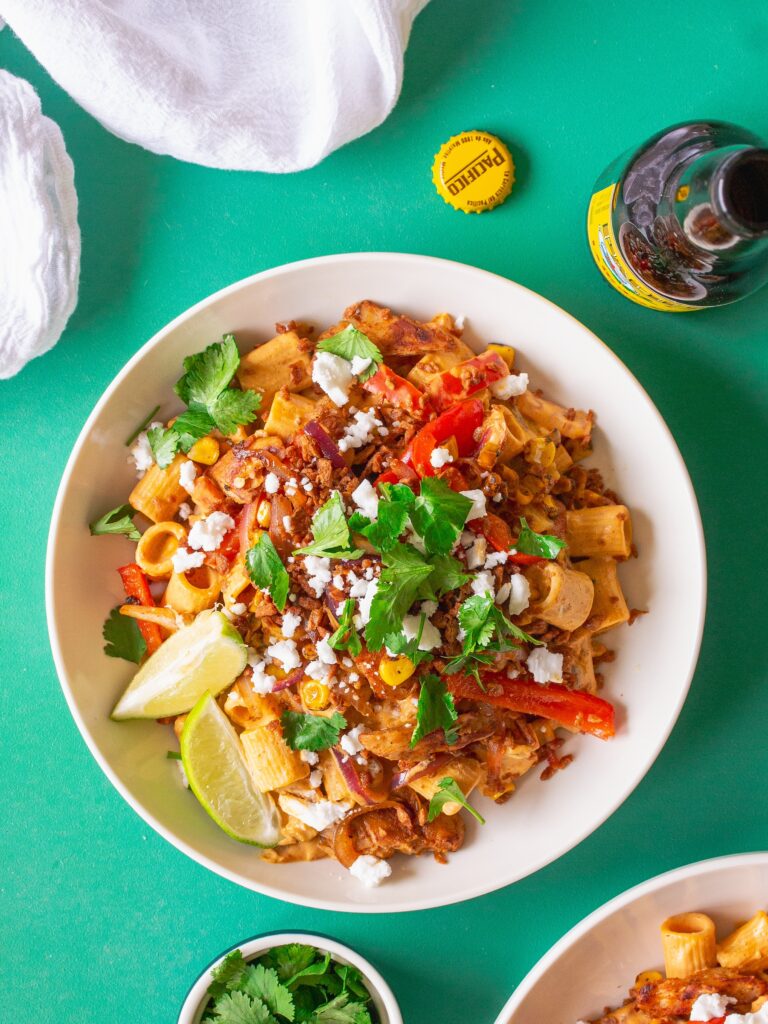 A serving plate showcasing the final dish of Vegan Chipotle Chicken Pasta, topped with vegan feta, bac'n bits, and fresh cilantro, with lime wedges on the side.