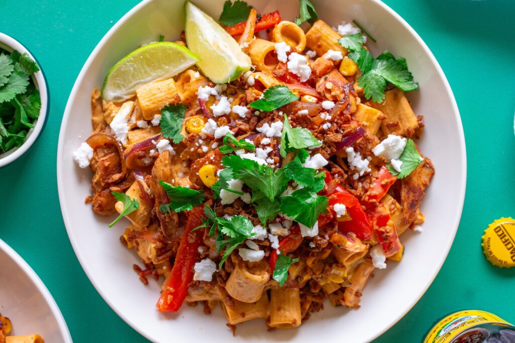 A serving plate showcasing the final dish of Vegan Chipotle Chicken Pasta, topped with vegan feta, bac'n bits, and fresh cilantro, with lime wedges on the side.