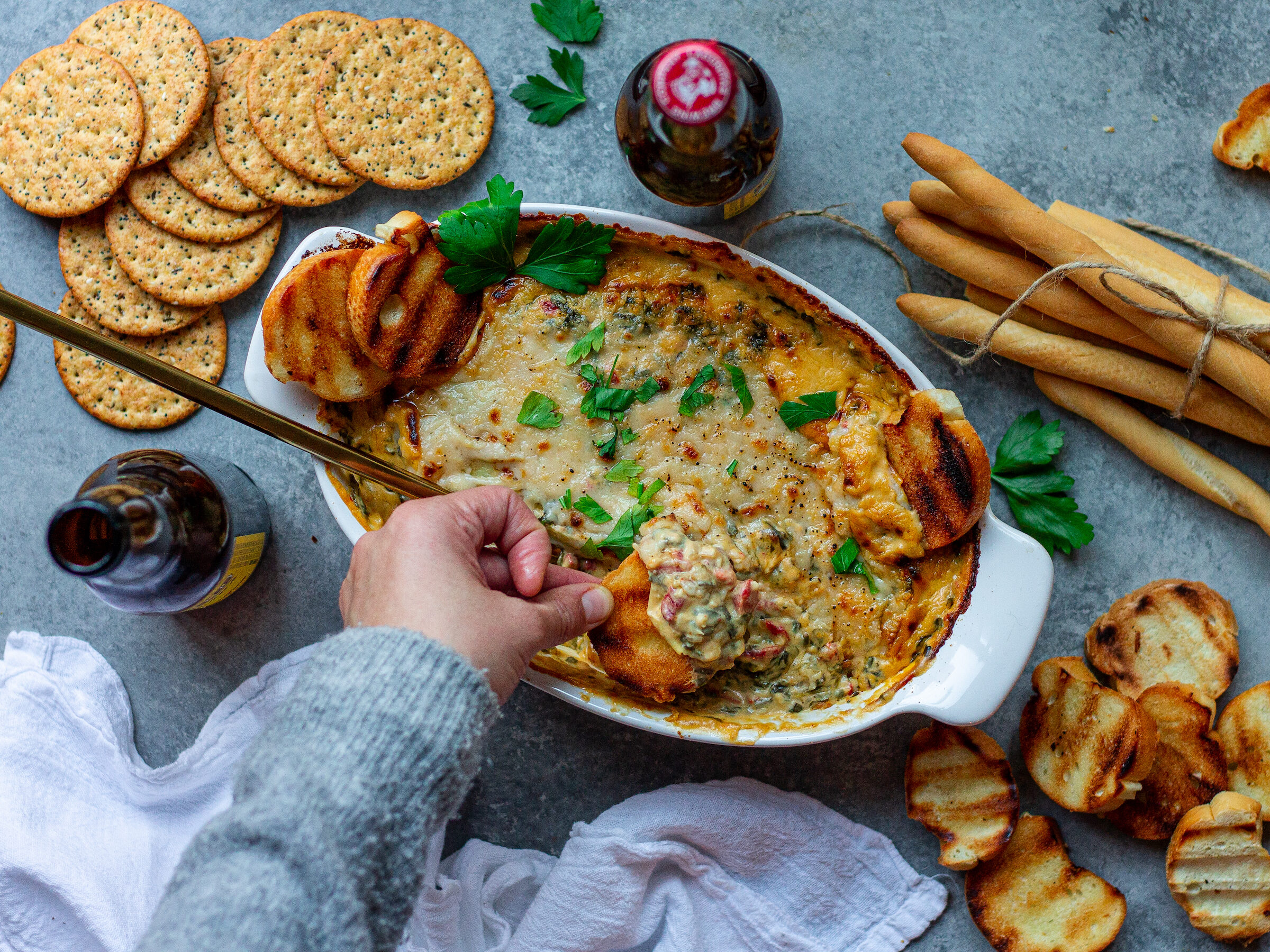 A creamy Vegan Spinach Artichoke Dip in a baking dish, topped with melted vegan parmesan and garnished with fresh parsley, surrounded by pita chips, toasted baguette slices, and veggies.