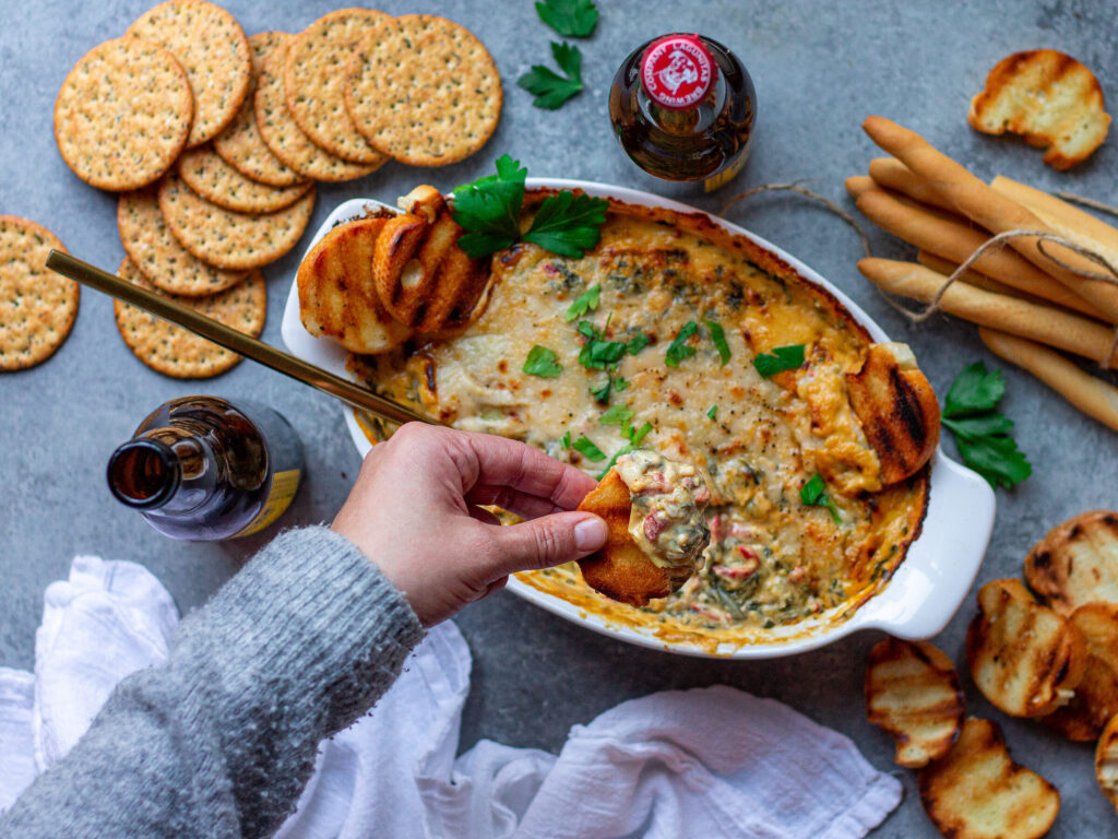 A hand dipping a toasted baguette slice into Vegan Spinach Artichoke Dip, showcasing its creamy and cheesy texture.