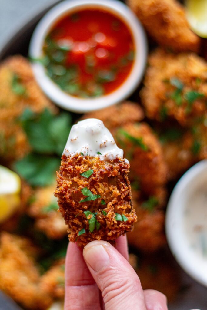 A hand dipping a crispy Parmesan Crusted Fried Artichoke Heart into a bowl of creamy vegan garlic mayo.