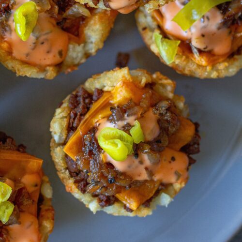 Golden tater tot cups filled with seasoned vegan ground beef, caramelized onions, melted vegan cheese, and topped with zesty burger sauce and banana peppers.