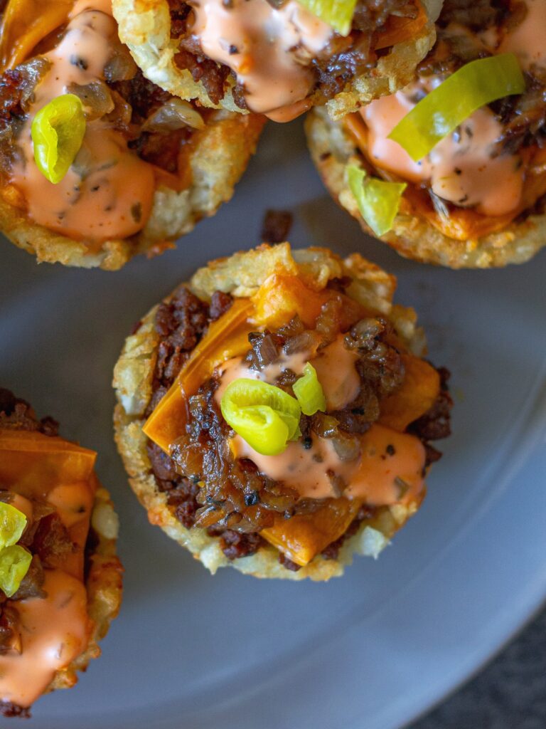 Golden tater tot cups filled with seasoned vegan ground beef, caramelized onions, melted vegan cheese, and topped with zesty burger sauce and banana peppers.