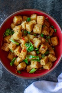 Crispy golden cubes of salt and pepper tofu garnished with green onions and Thai chilies on a serving plate.