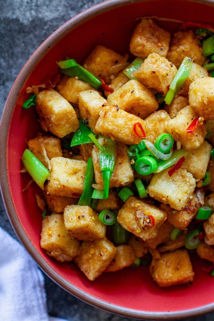 Crispy golden cubes of salt and pepper tofu garnished with green onions and Thai chilies on a serving plate.