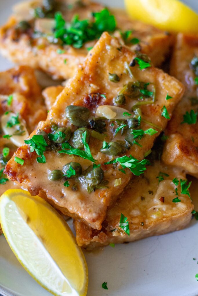 Detailed view of crispy tofu cutlets coated in tangy piccata sauce with visible capers and fresh herbs.
