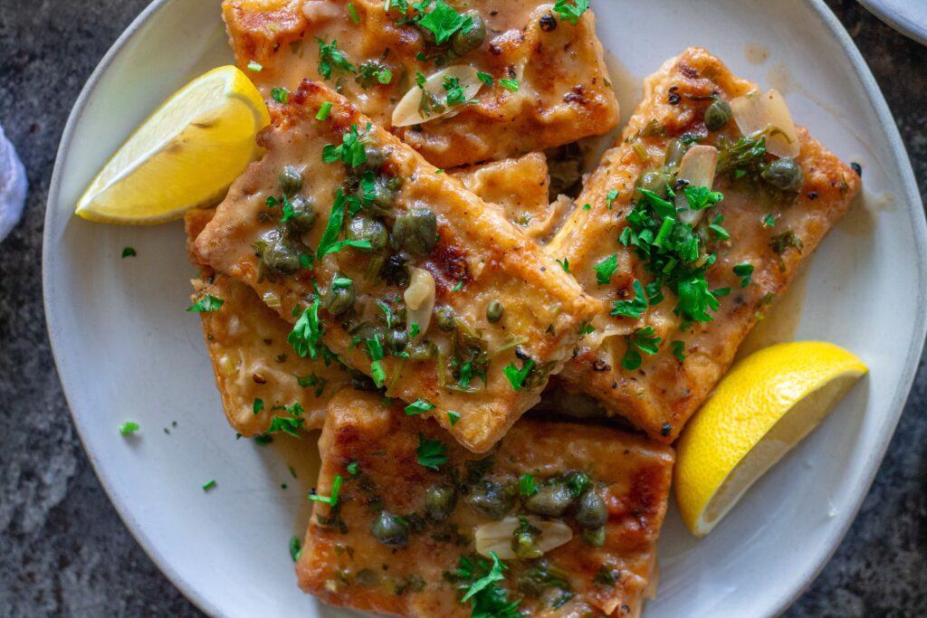 Tofu Piccata plated with mashed potatoes and a glass of white wine in the background for an elegant dining presentation.