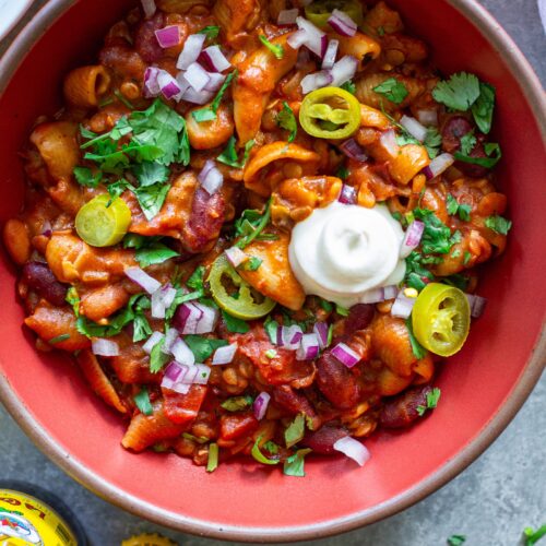 A bowl of cheesy and hearty One-Pot Lentil Chili Mac topped with fresh cilantro, red onion, pickled jalapeño, and vegan sour cream.