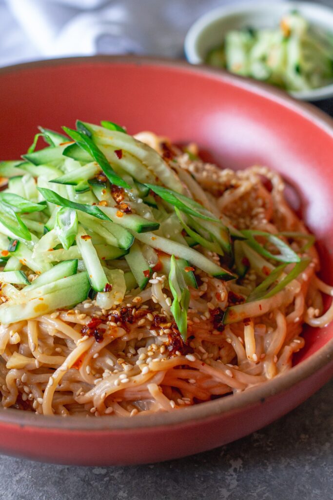 Close-up shot of cold sesame noodles in a bowl topped with sliced cucumbers and toasted sesame seeds.