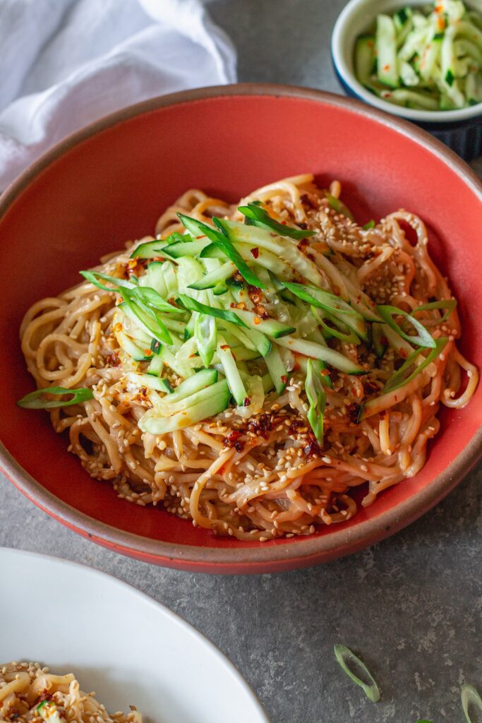 Close-up shot of cold sesame noodles in a bowl topped with sliced cucumbers and toasted sesame seeds.